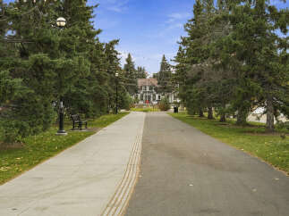 Discover this charming property featuring a picturesque pathway leading to a beautiful building, surrounded by lush green trees and a clear blue sky.