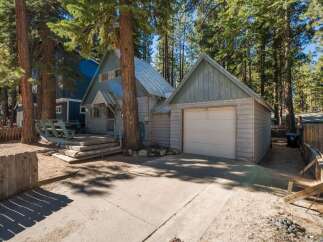 Nestled among tall pine trees, this charming property features a light blue A-frame house with a metal roof and a detached garage.