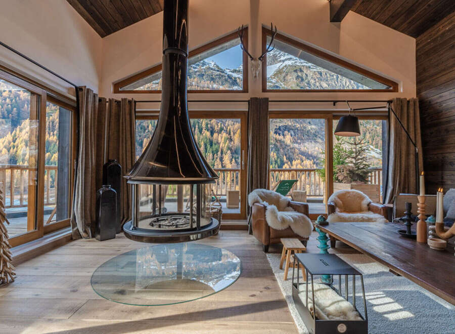 Experience the ultimate mountain retreat in this stunning living room, featuring a dramatic suspended fireplace and panoramic windows that showcase breathtaking autumnal forest and snow-capped peaks.
