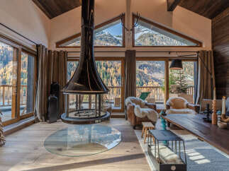 Experience the ultimate mountain retreat in this stunning living room, featuring a dramatic suspended fireplace and panoramic windows that showcase breathtaking autumnal forest and snow-capped peaks.