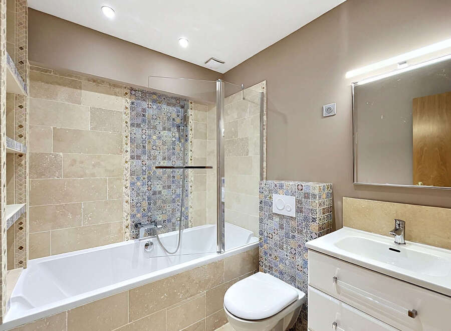 This beautifully appointed bathroom features a spacious bathtub with a glass shower screen, complemented by elegant travertine wall tiles and intricate mosaic accents.