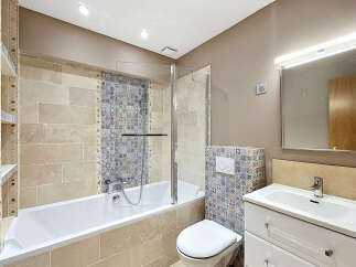 This beautifully appointed bathroom features a spacious bathtub with a glass shower screen, complemented by elegant travertine wall tiles and intricate mosaic accents.
