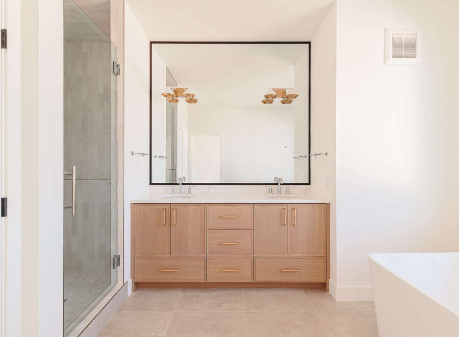 This modern bathroom features a spacious double vanity with light wood cabinetry and sleek rose gold hardware, complemented by a large frameless mirror.