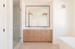 This modern bathroom features a spacious double vanity with light wood cabinetry and sleek rose gold hardware, complemented by a large frameless mirror.