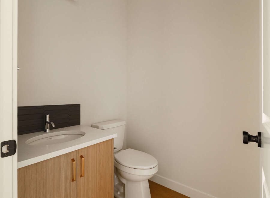 This modern powder room features a sleek vanity with a white countertop and warm wood cabinetry, complemented by a stylish dark tile backsplash.