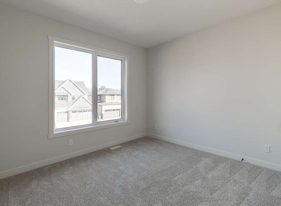 This empty room features a large window offering a view of suburban homes and a plush, neutral-toned carpet.