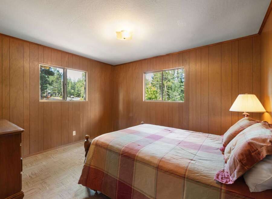 This cozy bedroom features warm wood-paneled walls and two large windows offering serene views of the surrounding pine forest.