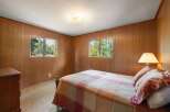 This cozy bedroom features warm wood-paneled walls and two large windows offering serene views of the surrounding pine forest.