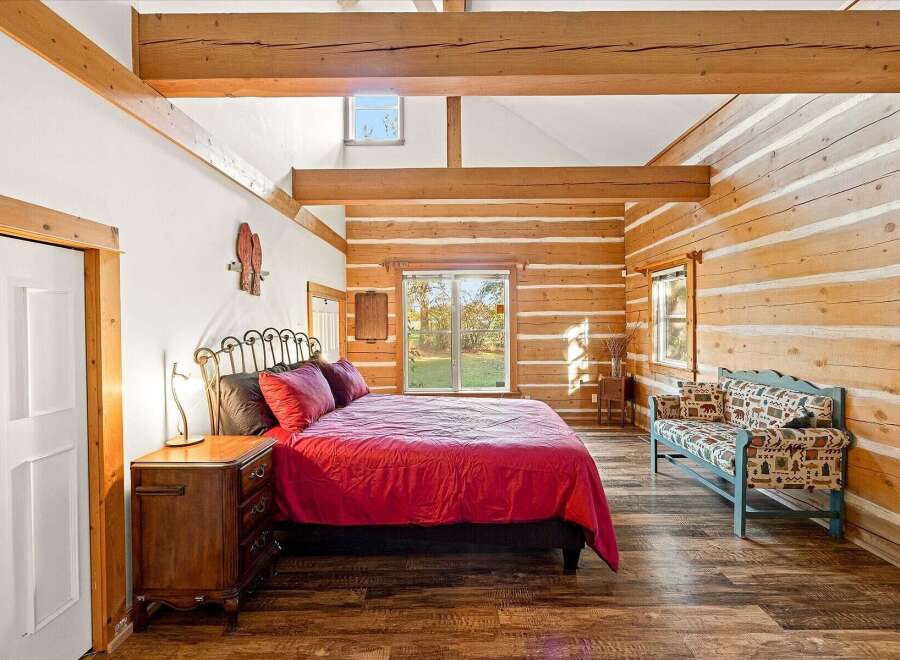 Experience rustic charm in this cozy log cabin bedroom, featuring a vibrant red comforter on a metal-frame bed and a charming patterned bench.