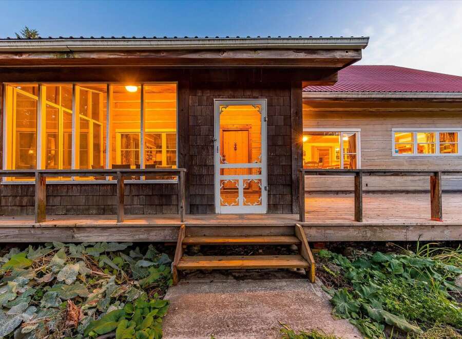 Step onto the inviting wooden porch of this charming home, featuring a rustic screened door and large windows that glow with warm interior light.