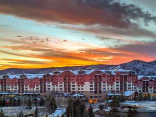 Experience luxury mountain living in this stunning ski resort property, featuring a grand red-accented building with snow-capped roofs and expansive windows.