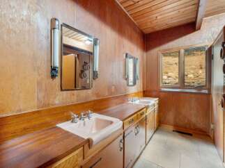 This rustic bathroom features a long wooden vanity with two pink sinks and chrome fixtures, complemented by wall-mounted light sconces and mirrors.