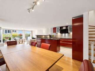 This modern open-concept living space features a spacious dining area with a wooden table and plush seating, seamlessly flowing into a sleek, red-accented kitchen.