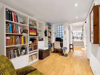 Discover a bright and organized living space featuring extensive white shelving filled with books and decorative items, complemented by a cozy armchair and a dedicated workspace.