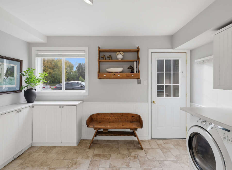 This charming laundry room features ample white cabinetry for storage, complemented by a large window offering natural light and a view of the outdoors.