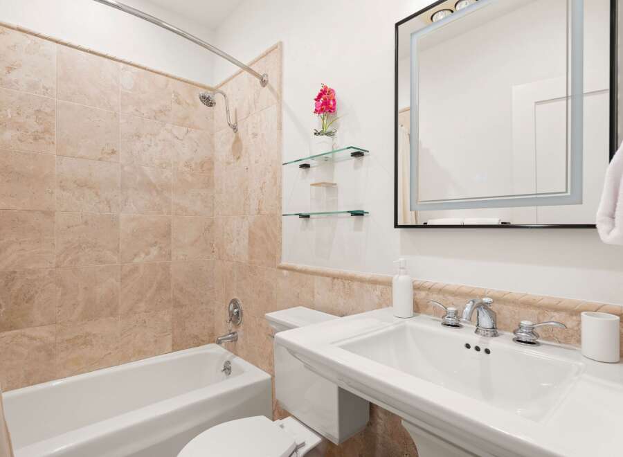This elegant bathroom features a luxurious bathtub with travertine tile surround, complemented by a sleek pedestal sink with chrome fixtures.