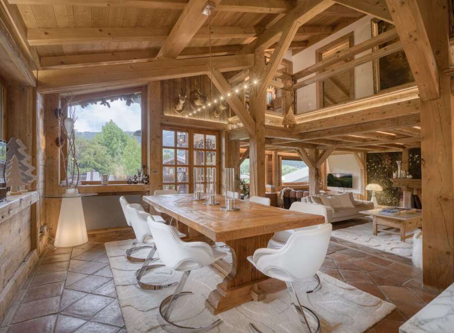 Experience luxurious mountain living in this stunning chalet, featuring a grand dining area with a rustic wooden table and modern white chairs, complemented by exposed wooden beams and a cozy fireplace.