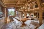 Experience luxurious mountain living in this stunning chalet, featuring a grand dining area with a rustic wooden table and modern white chairs, complemented by exposed wooden beams and a cozy fireplace.