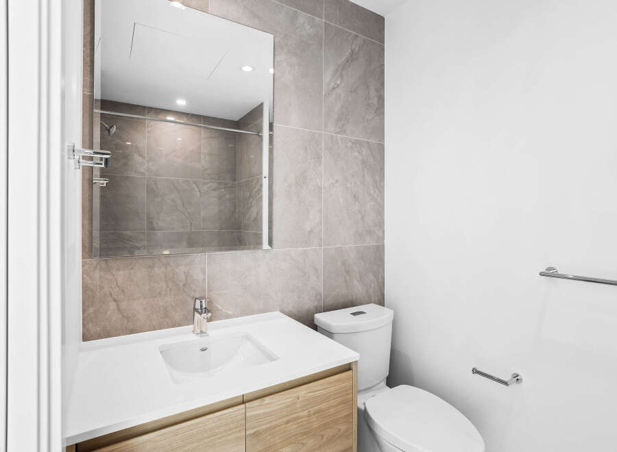 This modern bathroom features a sleek vanity with a white countertop and light wood cabinetry, complemented by a large, illuminated mirror.