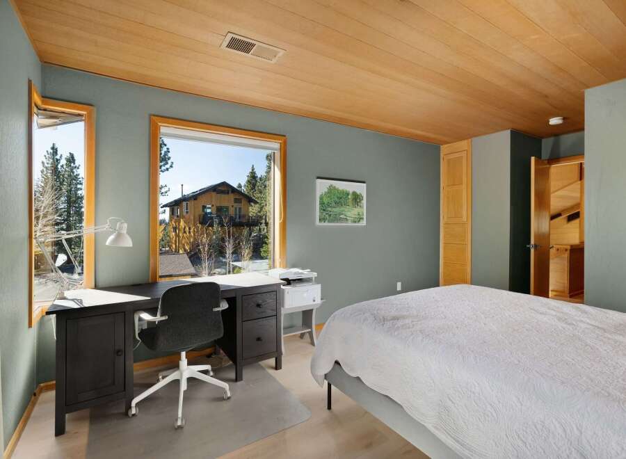 This bedroom offers a functional workspace with a large desk, an ergonomic chair, and a printer, all overlooking a scenic view of the surrounding nature and a charming house.