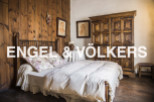 This charming bedroom boasts a rustic wooden wall, a cozy bed with crocheted details, and an antique wooden wardrobe.