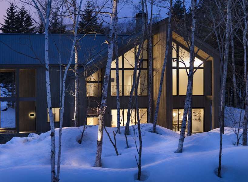 This stunning modern home nestled in a snowy forest boasts expansive windows showcasing breathtaking views, creating a warm and inviting atmosphere.
