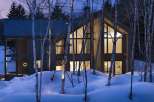 This stunning modern home nestled in a snowy forest boasts expansive windows showcasing breathtaking views, creating a warm and inviting atmosphere.