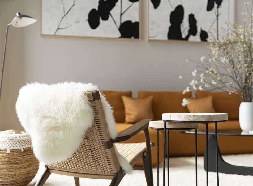 A cozy living room features a stylish woven chair with a sheepskin throw, complemented by a warm orange velvet sofa and minimalist side tables.