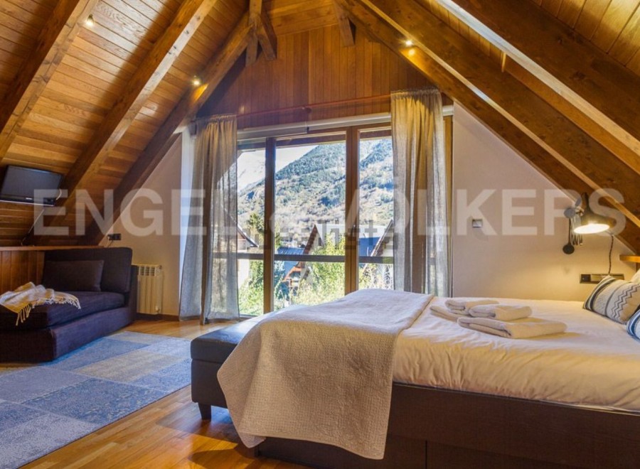 This stunning attic bedroom boasts a cozy wooden interior, large windows offering breathtaking mountain views, and a comfortable bed with plush bedding.