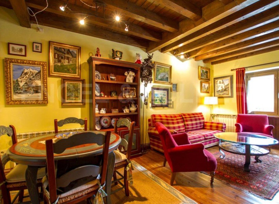 Charming apartment featuring exposed wooden beams, a cozy living area with plaid sofa and red armchairs, and a quaint dining area with a hand-painted table.