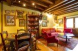 Charming apartment featuring exposed wooden beams, a cozy living area with plaid sofa and red armchairs, and a quaint dining area with a hand-painted table.