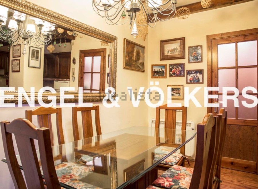Charming dining room with a glass-top table and wooden chairs, complemented by a large ornate mirror reflecting the kitchen and a beautiful chandelier.