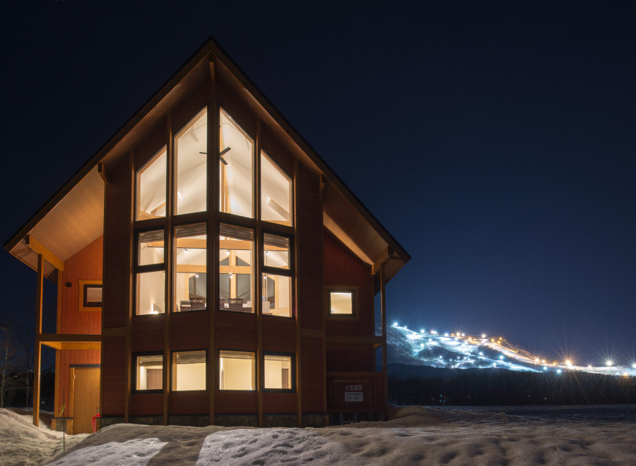 This stunning A-frame chalet boasts a wall of windows showcasing breathtaking nighttime views of a snow-covered mountain with illuminated ski slopes.