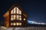 This stunning A-frame chalet boasts a wall of windows showcasing breathtaking nighttime views of a snow-covered mountain with illuminated ski slopes.