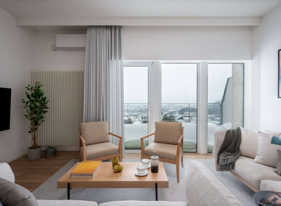 Modern living room with floor-to-ceiling windows offering stunning city views, featuring light beige sofas and armchairs, a wooden coffee table, and a minimalist design aesthetic.