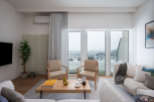 Modern living room with floor-to-ceiling windows offering stunning city views, featuring light beige sofas and armchairs, a wooden coffee table, and a minimalist design aesthetic.