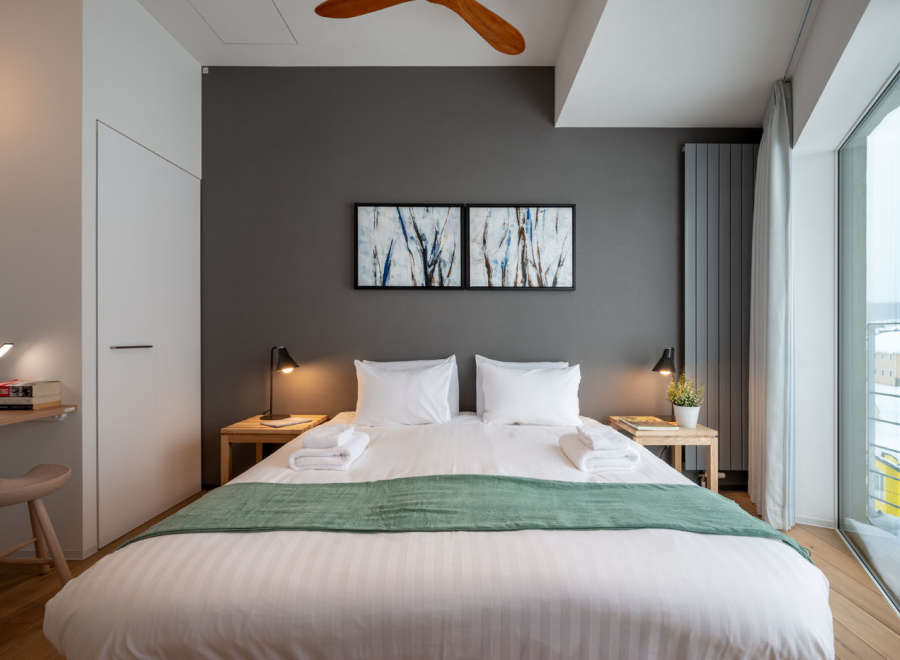 This modern bedroom boasts a king-size bed with crisp white linens and a plush green throw, complemented by minimalist nightstands and stylish artwork.