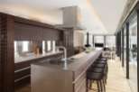 Modern kitchen with stainless steel countertops and dark wood cabinetry, featuring a large island with bar stools and a stunning view through floor-to-ceiling windows.