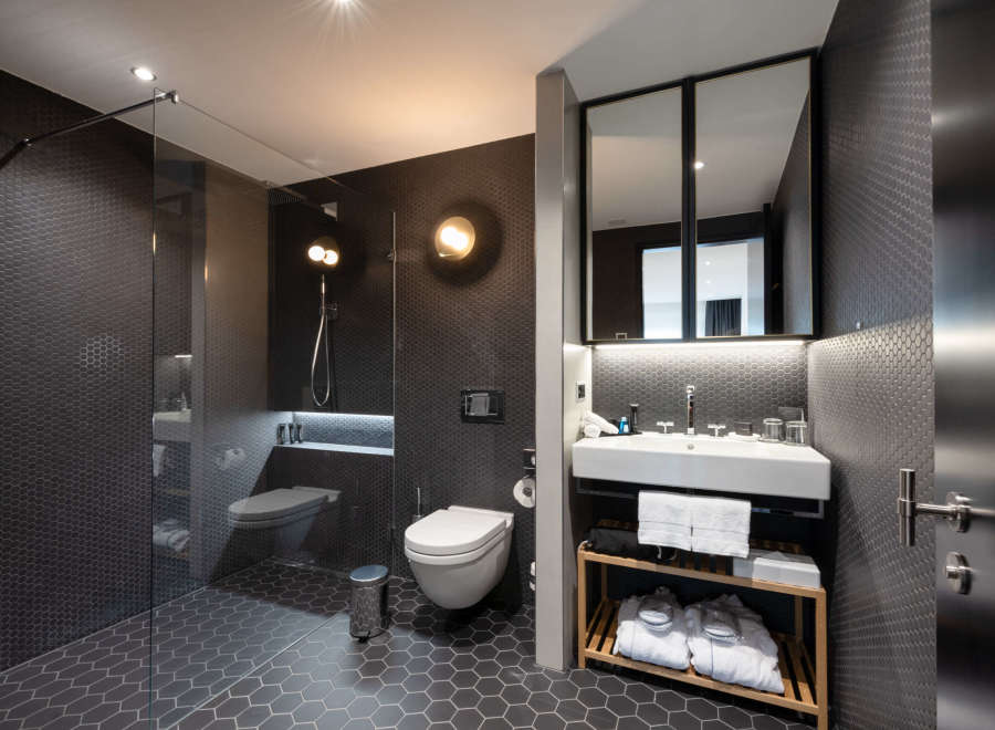 This modern bathroom boasts a sleek design with dark hexagon tiles, a glass shower enclosure, and a floating vanity with a wooden shelf for toiletries and towels.