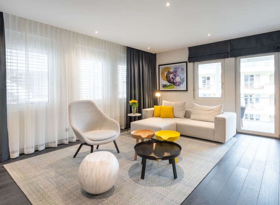 Modern living room with light beige couch and armchair, complemented by dark gray curtains and flooring.