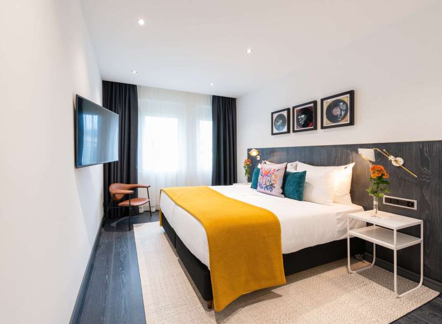 Stylish hotel room featuring a comfortable king-size bed with a mustard yellow throw, complemented by dark wood paneling and modern wall art.