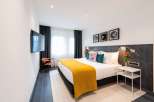 Stylish hotel room featuring a comfortable king-size bed with a mustard yellow throw, complemented by dark wood paneling and modern wall art.