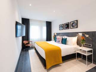 Stylish hotel room featuring a comfortable king-size bed with a mustard yellow throw, complemented by dark wood paneling and modern wall art.