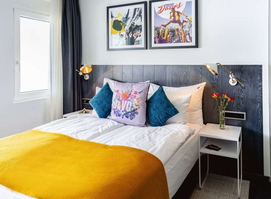 Stylish hotel room featuring a comfortable double bed with a mustard yellow throw, complemented by teal and floral patterned pillows.