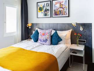 Stylish hotel room featuring a comfortable double bed with a mustard yellow throw, complemented by teal and floral patterned pillows.
