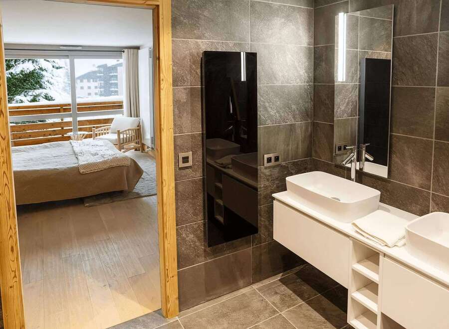 Luxurious bathroom with modern double sinks and heated towel rack, offering stunning views of a snowy mountain landscape from the adjacent bedroom.