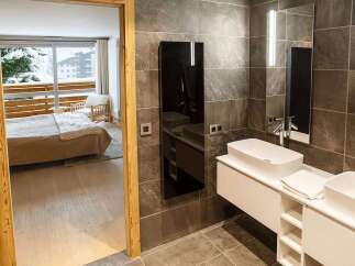 Luxurious bathroom with modern double sinks and heated towel rack, offering stunning views of a snowy mountain landscape from the adjacent bedroom.