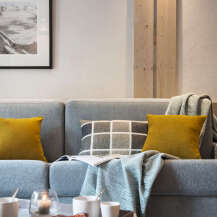 Cozy living room with a gray sofa featuring mustard yellow and gray patterned throw pillows, a soft teal throw blanket, and a framed mountain landscape print on the wall.