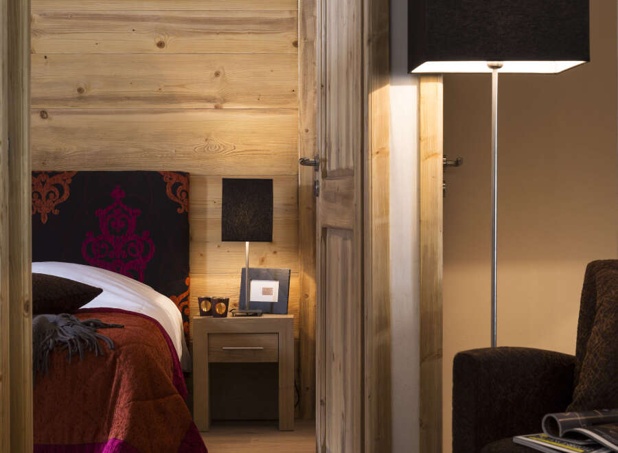 This cozy bedroom features a rustic wooden wall, a comfortable bed with a rich colored quilt, and a bedside table with a lamp.