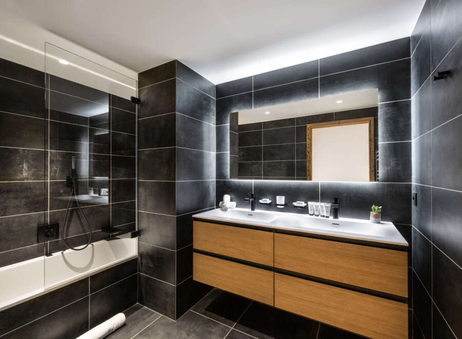 This luxurious bathroom boasts a sleek, modern design with dark gray tile, a spacious double vanity with light wood cabinets, and a frameless glass shower enclosure.